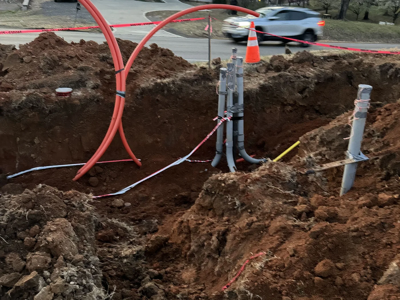 Open utility trench with orange conduit and gray pipe stubs in front of a house